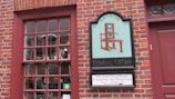 A brick wall features a window with a red frame and a multi-pane design. Next to it, a vintage-style sign displays an illustration of an old printing press, with the text 'Edes & Gill Printing Office 1755.' Below is an additional sign indicating a Colonial Print Shop demonstration at Old North Church.