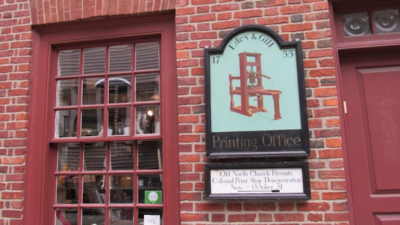 A brick wall features a window with a red frame and a multi-pane design. Next to it, a vintage-style sign displays an illustration of an old printing press, with the text 'Edes & Gill Printing Office 1755.' Below is an additional sign indicating a Colonial Print Shop demonstration at Old North Church.