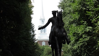 A silhouette of a horse and rider statue set against a backdrop of lush green trees, with a tall white church steeple visible in the distance.