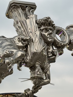 A highly reflective, metallic sculpture featuring an intricately designed human figure with curly hair, draped in flowing garments. The sculpture appears to be suspended or floating and exhibits an abstract fusion of classical and modern elements.