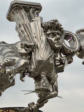 A highly reflective, metallic sculpture featuring an intricately designed human figure with curly hair, draped in flowing garments. The sculpture appears to be suspended or floating and exhibits an abstract fusion of classical and modern elements.