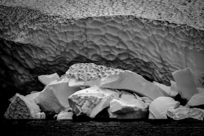 Layers of ice formations with intricate textures and patterns, showing large chunks of ice resting at the edge of a body of water. The surface appears rough and uneven, creating a dramatic, natural sculpture-like effect.