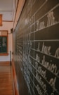 A classroom setting with a large blackboard displaying neatly written cursive handwriting, focusing on letters and phrases. The room is softly lit, with wooden flooring and trim visible. Another smaller board can be seen in the background.