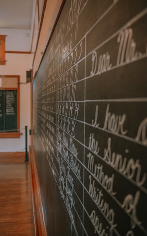 A classroom setting with a large blackboard displaying neatly written cursive handwriting, focusing on letters and phrases. The room is softly lit, with wooden flooring and trim visible. Another smaller board can be seen in the background.