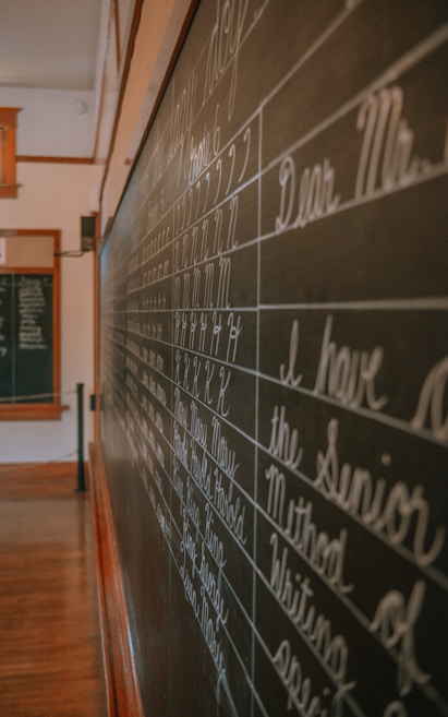 A classroom setting with a large blackboard displaying neatly written cursive handwriting, focusing on letters and phrases. The room is softly lit, with wooden flooring and trim visible. Another smaller board can be seen in the background.
