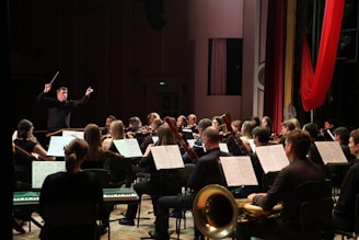 An orchestra performs in a concert hall, with musicians playing various instruments such as violins, bassoons, and trumpets. A conductor stands at the front, energetically directing the ensemble. Music stands with sheets are visible, and the setting is a formal stage with red drapery.
