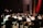 An orchestra performs in a concert hall, with musicians playing various instruments such as violins, bassoons, and trumpets. A conductor stands at the front, energetically directing the ensemble. Music stands with sheets are visible, and the setting is a formal stage with red drapery.