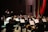 An orchestra performs in a concert hall, with musicians playing various instruments such as violins, bassoons, and trumpets. A conductor stands at the front, energetically directing the ensemble. Music stands with sheets are visible, and the setting is a formal stage with red drapery.