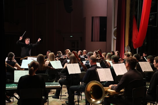 An orchestra performs in a concert hall, with musicians playing various instruments such as violins, bassoons, and trumpets. A conductor stands at the front, energetically directing the ensemble. Music stands with sheets are visible, and the setting is a formal stage with red drapery.