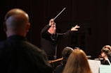 A conductor is energetically leading an orchestra, with musicians focused on their music sheets. The conductor is holding a baton in one hand, gesturing expressively. The lighting creates a dramatic atmosphere with highlights on the conductor and musicians.