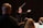 A conductor is energetically leading an orchestra, with musicians focused on their music sheets. The conductor is holding a baton in one hand, gesturing expressively. The lighting creates a dramatic atmosphere with highlights on the conductor and musicians.