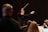 A conductor is energetically leading an orchestra, with musicians focused on their music sheets. The conductor is holding a baton in one hand, gesturing expressively. The lighting creates a dramatic atmosphere with highlights on the conductor and musicians.
