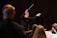 A close-up of a conductor passionately leading an orchestra in a dimly lit studio.