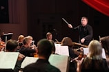 A conductor energetically leads an orchestra, surrounded by musicians playing violins and cellos. Sheet music is visible, suggesting a live performance in a dimly lit concert hall.
