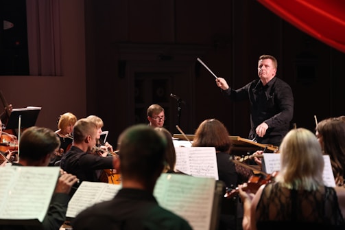 A conductor energetically leads an orchestra, surrounded by musicians playing violins and cellos. Sheet music is visible, suggesting a live performance in a dimly lit concert hall.