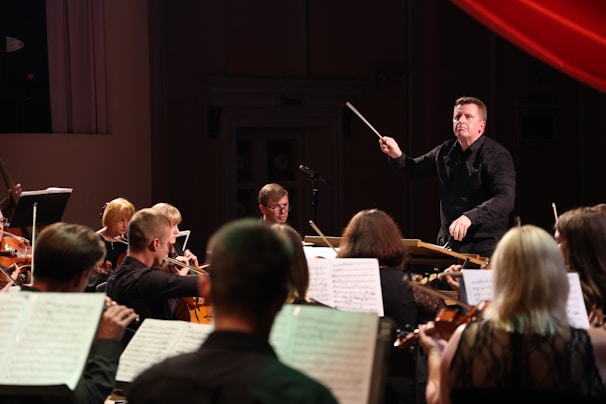 A conductor energetically leads an orchestra, surrounded by musicians playing violins and cellos. Sheet music is visible, suggesting a live performance in a dimly lit concert hall.