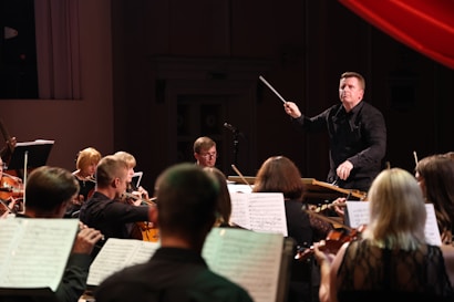 A conductor energetically leads an orchestra, surrounded by musicians playing violins and cellos. Sheet music is visible, suggesting a live performance in a dimly lit concert hall.