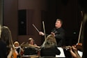 A conductor passionately leads an orchestra, with musicians attentively playing their string instruments. The setting is a concert hall with dim lighting focused on the performers.