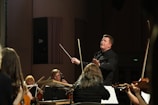A conductor passionately leads an orchestra, with musicians attentively playing their string instruments. The setting is a concert hall with dim lighting focused on the performers.