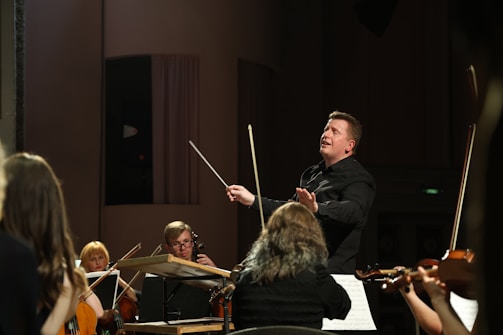 A conductor passionately leads an orchestra, with musicians attentively playing their string instruments. The setting is a concert hall with dim lighting focused on the performers.