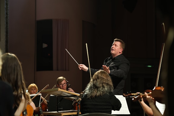 A conductor passionately leads an orchestra, with musicians attentively playing their string instruments. The setting is a concert hall with dim lighting focused on the performers.