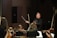 A conductor passionately leads an orchestra, with musicians attentively playing their string instruments. The setting is a concert hall with dim lighting focused on the performers.