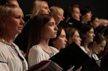 A group of individuals, mostly women, are gathered together, each holding a book or folder, possibly engaged in singing or a choir rehearsal. They are wearing coordinated white garments with dark embroidery. Their expressions are focused and attentive.