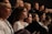 A group of individuals, mostly women, are gathered together, each holding a book or folder, possibly engaged in singing or a choir rehearsal. They are wearing coordinated white garments with dark embroidery. Their expressions are focused and attentive.