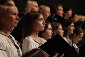 A group of individuals, mostly women, are gathered together, each holding a book or folder, possibly engaged in singing or a choir rehearsal. They are wearing coordinated white garments with dark embroidery. Their expressions are focused and attentive.