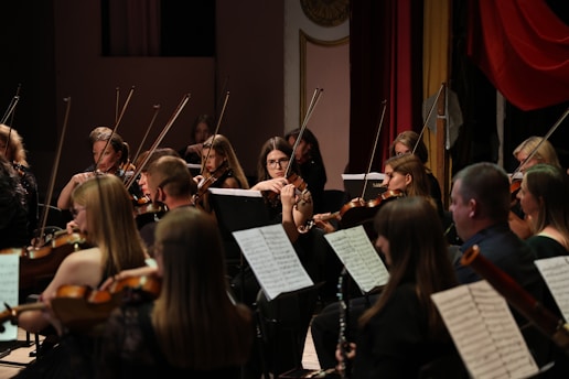 A group of musicians is playing in an orchestral setting. They are seated with various string instruments, including violins and violas, focused on their sheet music. The surrounding decor features a mixture of red and gold drapery, adding an elegant backdrop to the performance.