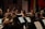 A group of musicians is playing in an orchestral setting. They are seated with various string instruments, including violins and violas, focused on their sheet music. The surrounding decor features a mixture of red and gold drapery, adding an elegant backdrop to the performance.