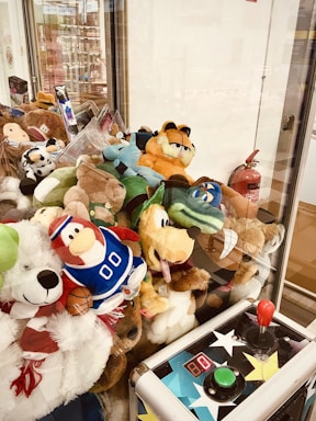 A claw machine filled with a variety of plush toys, including characters from cartoons and possibly comics. The control panel of the machine, with a joystick and buttons, is visible in the foreground. A fire extinguisher is mounted on the wall inside the glass encasing of the machine.