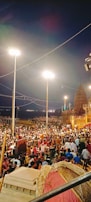 Wide shot of the riverfront filled with devotees watching the evening Ganga Aarti, illuminated by maroon and gold hues.
