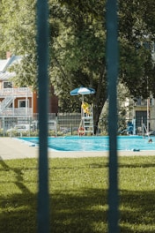 A public swimming pool is surrounded by a metal fence with a lifeguard in a yellow uniform sitting under a blue umbrella on a raised chair. The area features lush green grass and large trees, and the backdrop includes residential buildings.