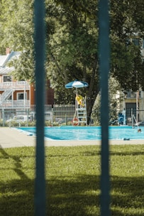A public swimming pool is surrounded by a metal fence with a lifeguard in a yellow uniform sitting under a blue umbrella on a raised chair. The area features lush green grass and large trees, and the backdrop includes residential buildings.