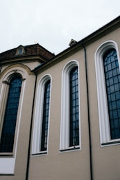 A section of an old building with tall, narrow arched windows. The structure features a beige facade with dark grid-patterned window panes. The architecture includes detailed molding and a tiled roof, suggesting historical or classical design influences.