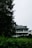 A house with a traditional design featuring a slate roof and multiple windows partially obscured by lush greenery. The building is nestled among trees and bushes, creating a serene and secluded atmosphere.