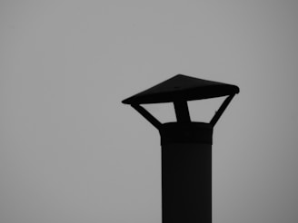 A silhouetted metal chimney cap with a pyramidal top sits against a plain gray background. Its geometric shape is distinct, with straight lines and a defined outline.