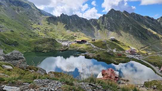 A picturesque mountainous landscape featuring a serene lake reflecting the sky and surrounding hills. The foreground contains rocky terrain with sparse grass, while the background showcases rugged mountains with grassy slopes. Several small buildings and structures are scattered around the lakeside, indicating a remote settlement or resort area.