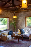 A cozy room with rustic walls and large windows featuring greenery outside. A plant is placed in the corner next to two white couches, covered with sheets and adorned with yellow pillows. A wooden coffee table holds books, a bottle, and other small items, while a patterned rug lies beneath. Two backpacks rest on the couches. Hanging lights cast a warm glow.