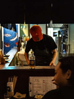 A dimly lit restaurant bar features a person wearing a red cap and apron, focused on preparing something behind the counter. A bottle stands prominently on the counter, surrounded by utensils and a menu booklet in the foreground. The overall ambiance is cozy and intimate.
