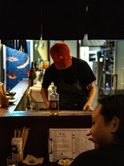 A dimly lit restaurant bar features a person wearing a red cap and apron, focused on preparing something behind the counter. A bottle stands prominently on the counter, surrounded by utensils and a menu booklet in the foreground. The overall ambiance is cozy and intimate.