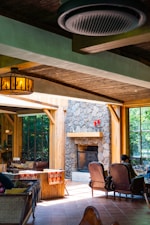 A cozy indoor space with wooden beams and a rustic, stone fireplace. Large windows allow natural light to flood the room, highlighting the warm wooden furnishings. Comfortable chairs and blankets suggest a welcoming and relaxing atmosphere. The room is adorned with a decorative light fixture and features a person sitting near the fireplace, adding to the homely vibe.