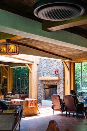 A cozy indoor space with wooden beams and a rustic, stone fireplace. Large windows allow natural light to flood the room, highlighting the warm wooden furnishings. Comfortable chairs and blankets suggest a welcoming and relaxing atmosphere. The room is adorned with a decorative light fixture and features a person sitting near the fireplace, adding to the homely vibe.