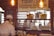 A cozy bakery or café scene with a brick wall interior and warm lighting. A menu board lists soups, including mushroom truffle and tomato rosée. A worker, wearing a white cap and mask, stands behind a counter with baked goods, such as bagels and bread. The shelves behind display plates, baskets, and stacked trays of eggs.