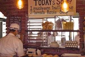 A cozy bakery or café scene with a brick wall interior and warm lighting. A menu board lists soups, including mushroom truffle and tomato rosée. A worker, wearing a white cap and mask, stands behind a counter with baked goods, such as bagels and bread. The shelves behind display plates, baskets, and stacked trays of eggs.