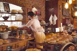 A person is working behind a bakery counter, surrounded by various types of bread and pastries. The setting features a rustic interior with brick walls and exposed light bulbs. Shelves are lined with plates and a picture is displayed on the wall. Some of the pastries are decorated with small British flags.
