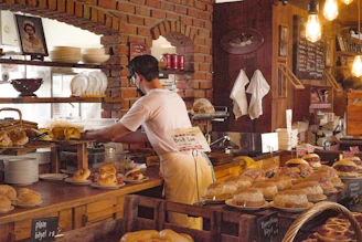 A person is working behind a bakery counter, surrounded by various types of bread and pastries. The setting features a rustic interior with brick walls and exposed light bulbs. Shelves are lined with plates and a picture is displayed on the wall. Some of the pastries are decorated with small British flags.
