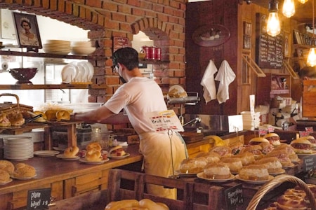 A person is working behind a bakery counter, surrounded by various types of bread and pastries. The setting features a rustic interior with brick walls and exposed light bulbs. Shelves are lined with plates and a picture is displayed on the wall. Some of the pastries are decorated with small British flags.