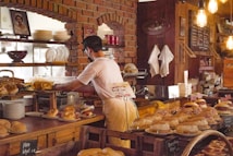 A person is working behind a bakery counter, surrounded by various types of bread and pastries. The setting features a rustic interior with brick walls and exposed light bulbs. Shelves are lined with plates and a picture is displayed on the wall. Some of the pastries are decorated with small British flags.
